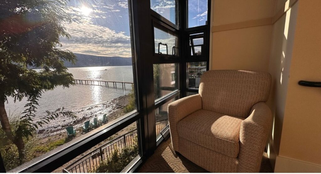 Beige upholstered armchair tucked into a sunlit corner beside floor to ceiling windows at Summerland Waterfront Resort and Spa. The windows overlook turquoise Adirondack chairs on the rocky lakeshore and a long dock extending across sparkling water.