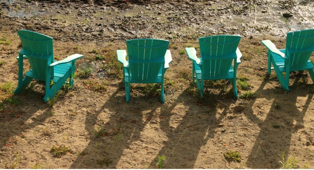 Four turquoise Adirondack chairs arranged in a row facing the rocky lakeshore at Summerland Waterfront Resort and Spa. Bright afternoon sunlight casts long chair shaped shadows across the sandy ground.