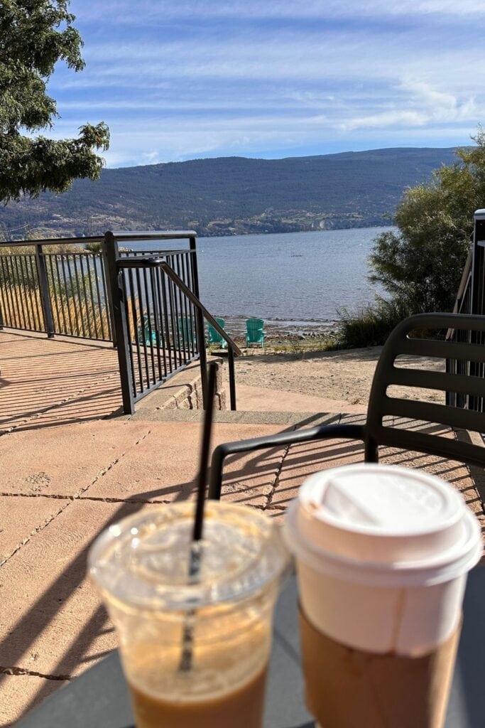 An iced coffee and a takeaway hot coffee sit on a patio table overlooking the waterfront at Summerland Waterfront Resort and Spa. A staircase leads down to turquoise Adirondack chairs beside the lake with mountains in the distance.