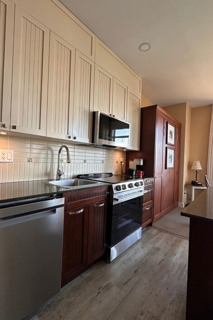 Fully equipped kitchenette at Summerland Waterfront Resort and Spa featuring cream upper cabinets and dark wooden lower cabinets. Stainless steel appliances include a dishwasher, oven, microwave and sink alongside a black countertop and white tile backsplash.