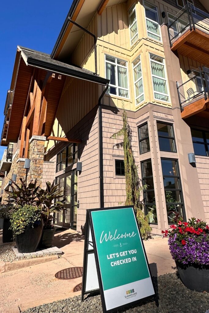 Exterior of Summerland Waterfront Resort and Spa featuring pale yellow siding, stone pillars, timber beams, large windows and glass balcony railings. A turquoise welcome sign outside the entrance reads "Welcome. LET'S GET YOU CHECKED IN."