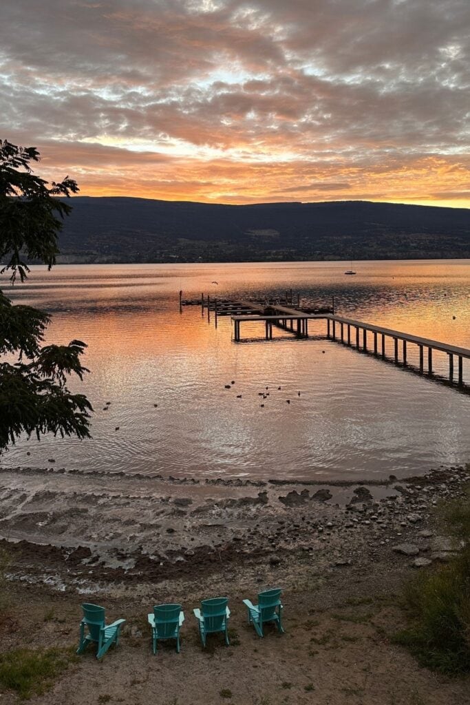 Orange and pink sunset reflecting across the lake beside Summerland Waterfront Resort and Spa. A long wooden dock extends into the water while four turquoise Adirondack chairs sit along the rocky shoreline beneath a dramatic cloud filled sky.
