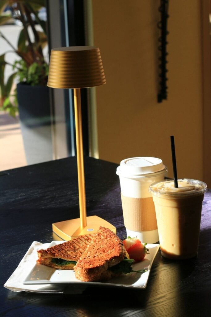 A toasted sandwich with leafy greens and a strawberry served on a white plate beside an iced coffee and a hot takeaway coffee. A small gold table lamp illuminates the breakfast spread on a dark wooden cafe table.