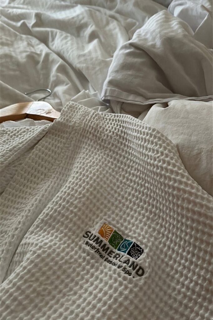 Two white waffle textured spa robes laid across a bed with rumpled white bedding and a wooden clothes hanger. One robe features an embroidered logo reading "SUMMERLAND Waterfront Resort and Spa" beneath four colourful squares.