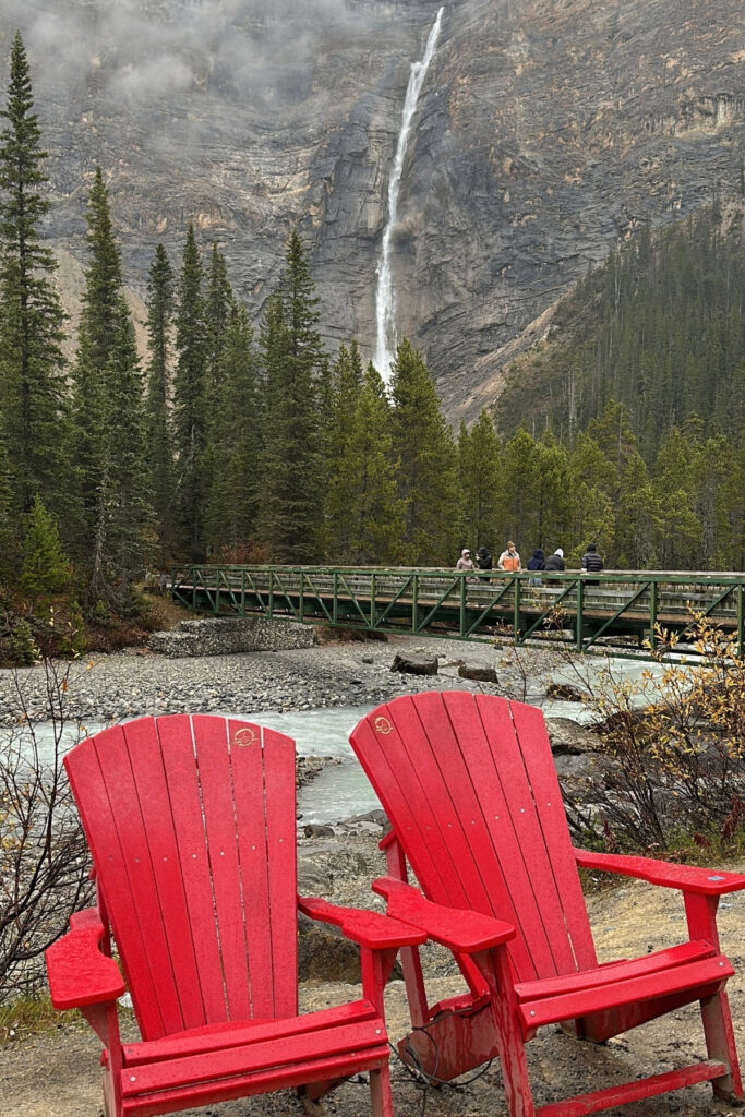Two red Adirondack chairs overlooking Takakkaw Falls in Yoho National Park, with a pedestrian bridge, evergreen forest and towering rock cliffs.